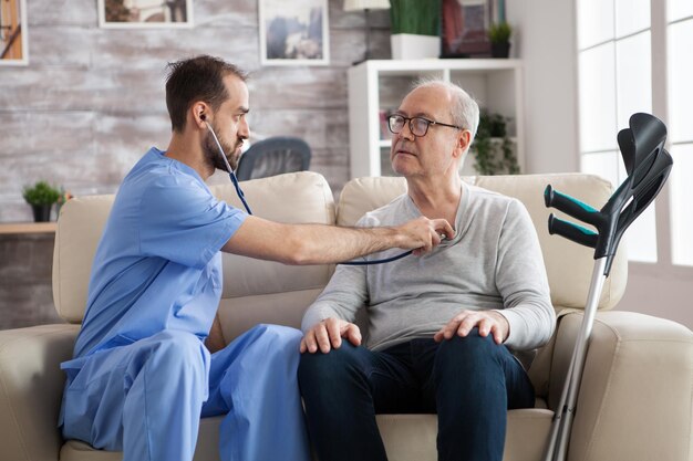 Junge männliche Krankenschwester mit Stethoskop, die das Herz eines älteren Mannes im Pflegeheim hört.