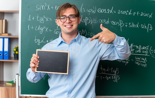Junge männliche Lehrer mit Brille, die eine kleine Tafel hält, die mit dem Zeigefinger darauf zeigt und lächelnd selbstbewusst in der Nähe einer Tafel mit mathematischen Formeln im Klassenzimmer steht