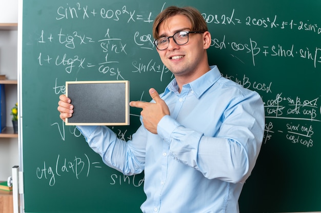 Junge männliche Lehrer mit Brille, die eine kleine Tafel hält und mit dem Zeigefinger darauf zeigt, lächelt selbstbewusst in der Nähe der Tafel mit mathematischen Formeln im Klassenzimmer