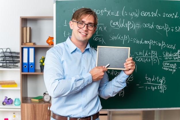 Kostenloses Foto junge männliche lehrer mit brille, die eine kleine tafel zeigt, die selbstbewusst in der nähe einer tafel mit mathematischen formeln im klassenzimmer lächelt