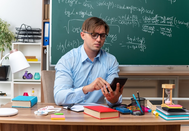 Junge männliche Lehrer mit Brille, die eine Nachricht mit dem Smartphone eingeben und selbstbewusst auf der Schulbank sitzen, mit Büchern und Notizen vor der Tafel im Klassenzimmer
