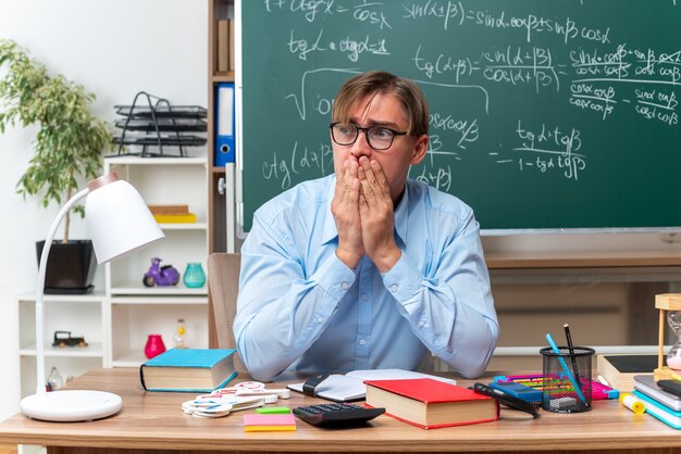 Junge männliche Lehrer mit Brille, die gestresst und nervös den Mund mit den Händen auf der Schulbank mit Büchern und Notizen vor der Tafel im Klassenzimmer bedecken