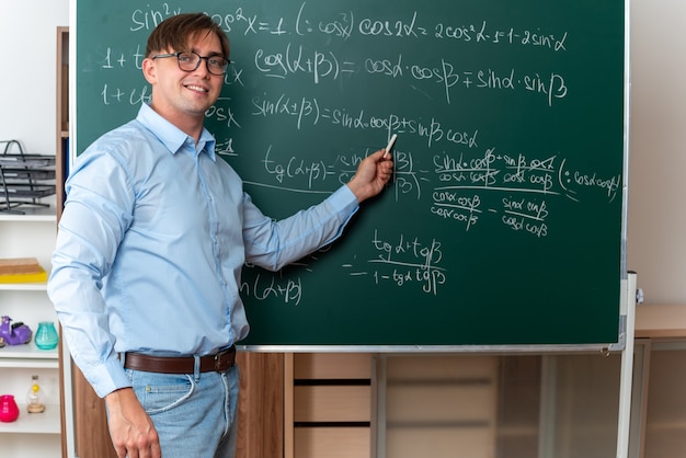 Junge männliche Lehrer mit Brille, die Kreide hält und die Lektion erklärt, lächelt selbstbewusst in der Nähe der Tafel mit mathematischen Formeln im Klassenzimmer