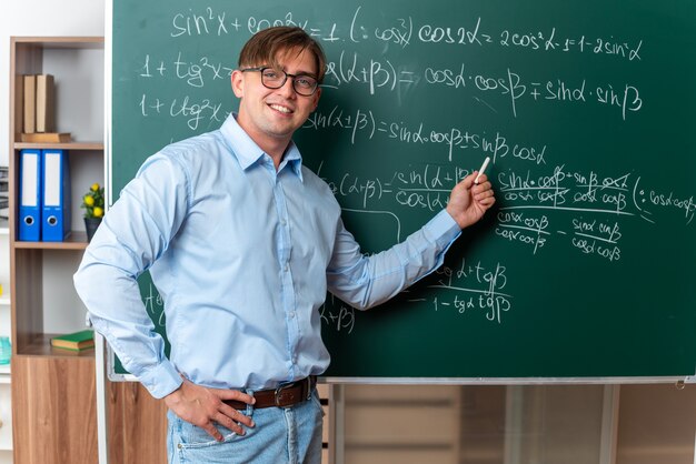 Junge männliche Lehrer mit Brille, die Kreide hält und die Lektion in der Nähe der Tafel mit mathematischen Formeln im Klassenzimmer erklärt