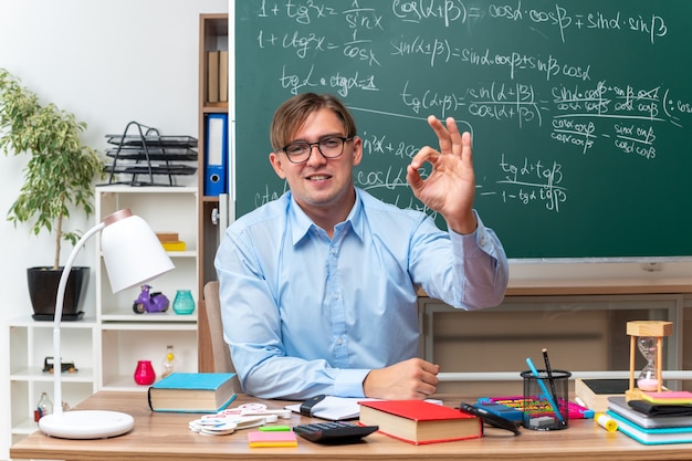 Kostenloses Foto junge männliche lehrer mit brille, die selbstbewusst lächeln und ein ok-zeichen auf der schulbank mit büchern und notizen vor der tafel im klassenzimmer zeigen