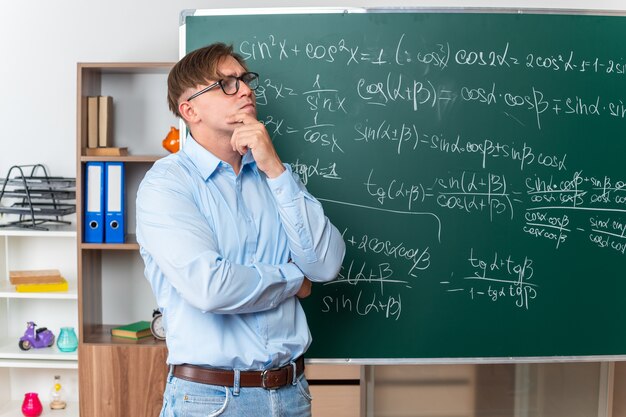 Junge männliche Lehrer mit Brille, die verwirrt in der Nähe der Tafel mit mathematischen Formeln im Klassenzimmer nachschlagen