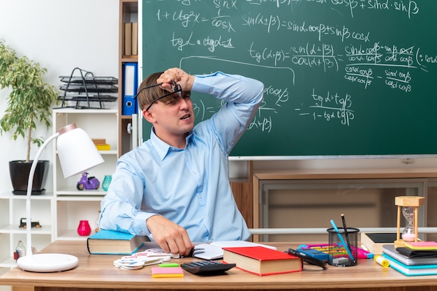 Kostenloses Foto junge männliche lehrer mit brille, die verwirrt und sehr ängstlich auf der schulbank mit büchern und notizen vor der tafel im klassenzimmer sitzen