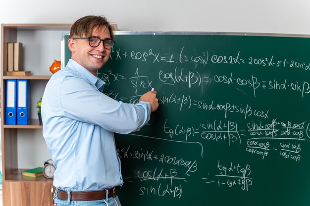 Junge männliche Lehrer mit Brille erklären die Lektion glücklich und positiv lächelnd in der Nähe der Tafel mit mathematischen Formeln im Klassenzimmer