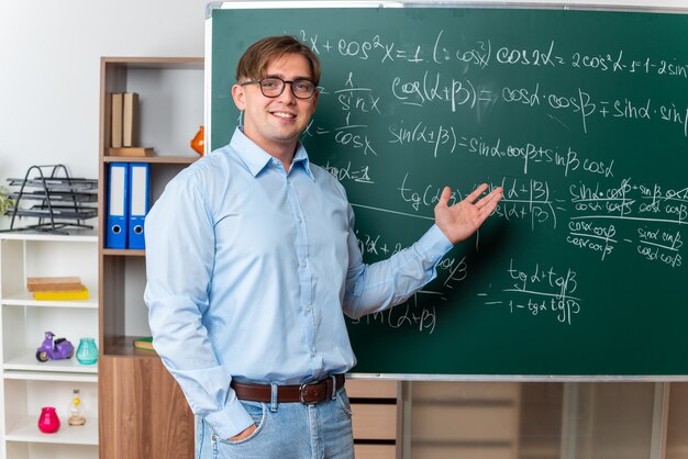 Junge männliche Lehrer mit Brille glücklich und positiv, die den Unterricht in der Nähe der Tafel mit mathematischen Formeln im Klassenzimmer erklären