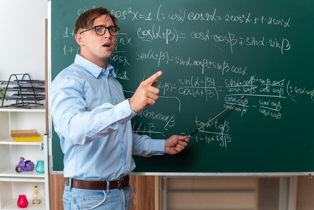 Junge männliche Lehrer mit Brille mit Zeiger, der die Lektion erklärt, die selbstbewusst in der Nähe der Tafel mit mathematischen Formeln im Klassenzimmer steht