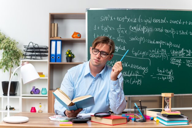 Junge männliche Lehrer mit Brille sitzen an der Schulbank mit Büchern und Notizen lesen Buch mit ernstem Gesicht vor der Tafel im Klassenzimmer