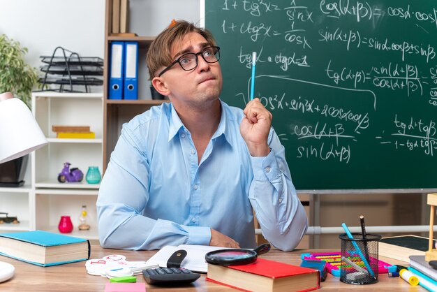 Junge männliche lehrer mit brille sitzen an der schulbank mit büchern und notizen mit bleistift, die verwirrt vor der tafel im klassenzimmer aussehen