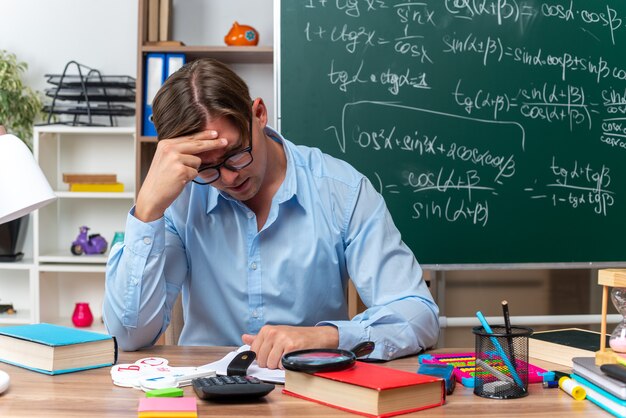 Junge männliche Lehrer mit Brille sitzen an der Schulbank mit Büchern und Notizen müde und gelangweilt vor der Tafel im Klassenzimmer