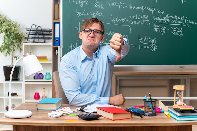Junge männliche Lehrer mit Brille unzufrieden mit Daumen nach unten sitzend an der Schulbank mit Büchern und Notizen vor der Tafel im Klassenzimmer