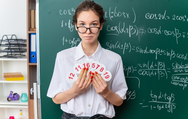 Junge mathematiklehrerin mit brille, die vor der tafel steht und zahlenfans hält, die im klassenzimmer nach vorne schauen?