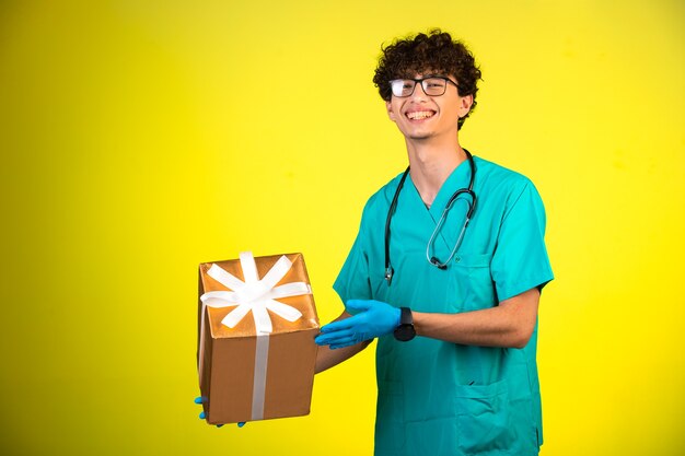 Junge mit lockigem Haar in medizinischer Uniform und Handmasken, die eine Geschenkbox halten.