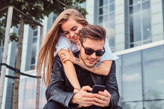 Junge modische Paare, die sich zusammen in der Nähe von Wolkenkratzern entspannen - hübsches Mädchen, das ihren Freund kuschelt, während er sein Smartphone benutzt.