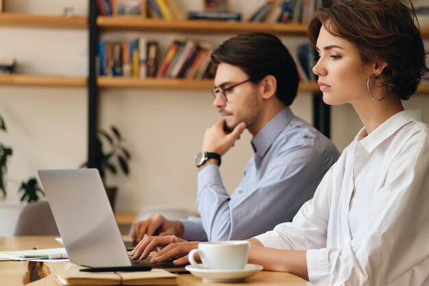 Junge müde Frau, die nachdenklich am Tisch sitzt und mit einem Kollegen in der Nähe eines modernen Büros am Laptop arbeitet