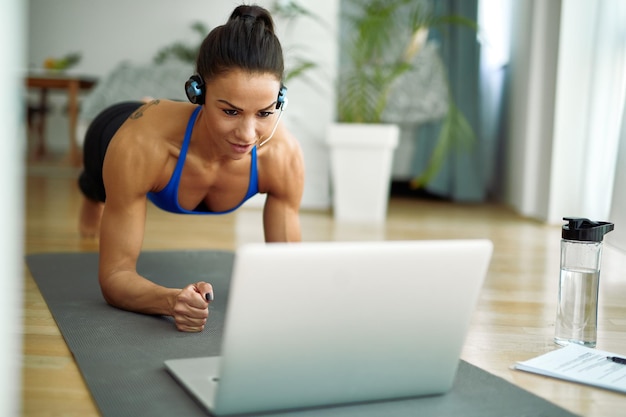 Junge muskulöse Frau, die in Plankenhaltung trainiert, während sie den Computer im Wohnzimmer benutzt
