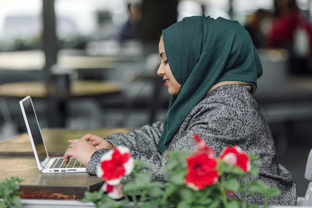 Junge muslimische Frau sitzt in einem Straßencafé und schaut in einen Laptop