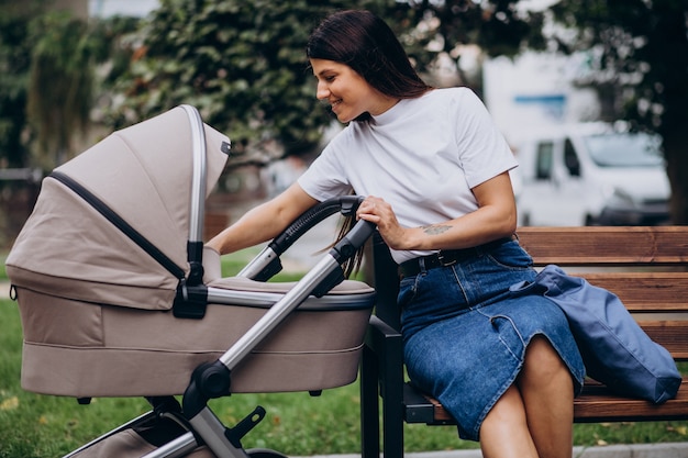 Junge Mutter, die auf Bank im Park mit Kinderwagen sitzt