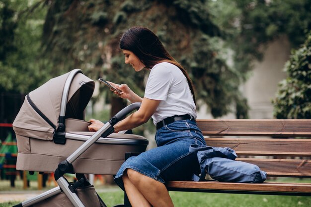 Junge Mutter, die auf Bank im Park mit Kinderwagen sitzt