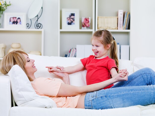 Junge Mutter, die auf dem Sofa liegt und mit ihrer glücklich liebenden kleinen Tochter spielt - drinnen