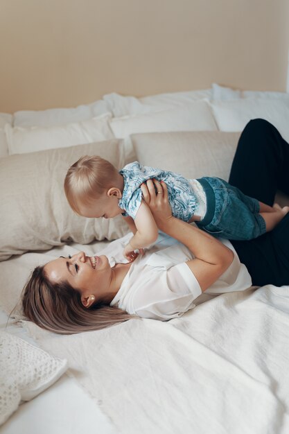 Junge Mutter, die Spaß am Lachen hat, das lustige aktive Spiele mit niedlichem Kindersohn im Schlafzimmer spielt