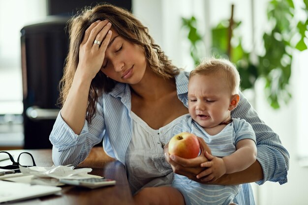 Junge Mutter fühlt sich erschöpft, während sie mit ihrem Baby zusammen ist und zu Hause arbeitet