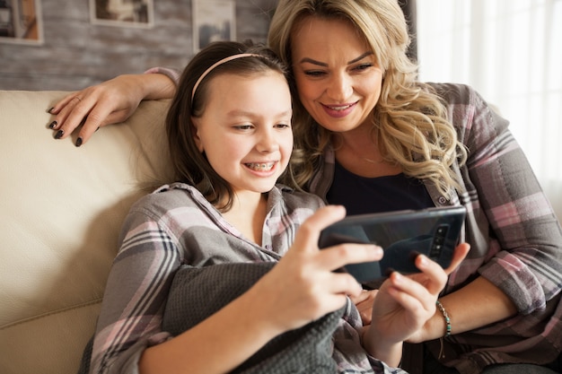 Junge Mutter lernt ihre kleine Tochter mit Klammern, um Anwendungen auf dem Smartphone zu verwenden, das auf der Couch im Wohnzimmer sitzt.