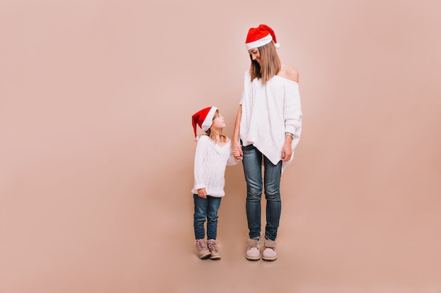 Junge Mutter mit der kleinen hübschen Tochter, die auf beige Wand steht und weiße Pullover und Weihnachtsmannmützen trägt, die einander schauen und Hände halten