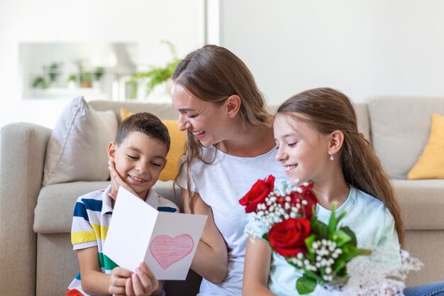 Junge Mutter mit einem Strauß Rosen lacht und umarmt ihren Sohn und ein fröhliches Mädchen mit einer Karte gratuliert Mutter während der Feiertagsfeier in der Küche zu Hause
