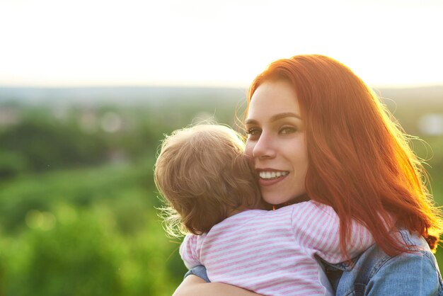 Junge Mutter umarmt Kind lächelnd auf schönem Panoramablick hinter Frau sieht glücklich aus, wenn sie Familienzeit mit Tochter lacht, die rote Haare trägt und blaues Jeanshemd trägt