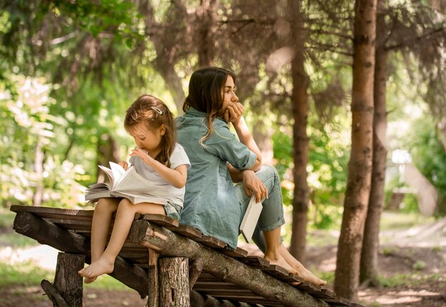 Junge Mutter und Tochter lasen im Park auf einer Holzbrücke ein Buch, das Konzept eines glücklichen Familienlebens und familiärer Beziehungen