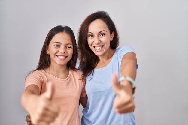 Junge Mutter und Tochter stehen vor weißem Hintergrund und machen zustimmend eine positive Geste mit erhobenem Daumen, lächeln und freuen sich über den Erfolg