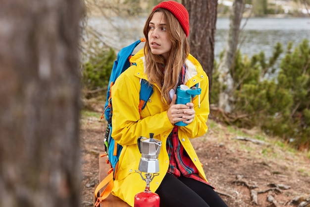 Junge nachdenkliche Frau mit Rucksack sitzt in einem kleinen Wald in der Nähe von Fluss oder See, wärmt sich mit heißem Getränk aus der Thermoskanne, kocht Kaffee auf Campingkocher