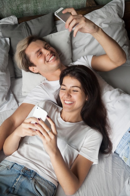 Junge Paare des Gadget-Süchtigen, die Spaß mit Smartphones im Bett haben