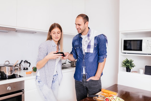 Junge Paare, die an der Küche unter Verwendung des Handys beim Kochen des Lebensmittels stehen