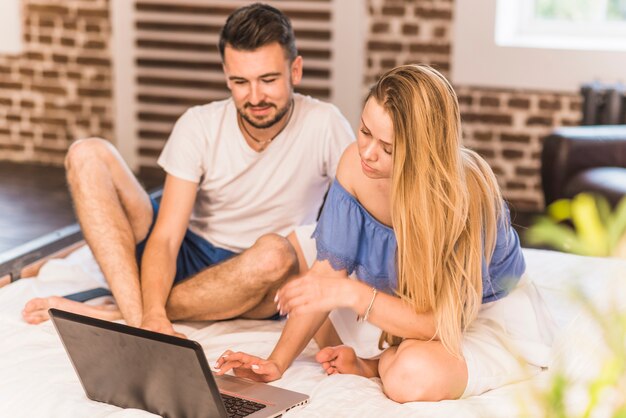 Junge Paare, die auf Bett unter Verwendung des Laptops sitzen