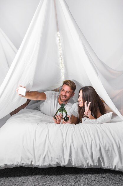 Junge Paare, die auf dem Bett nimmt Selfie auf dem Mobiltelefon hält Alkoholflaschen liegen