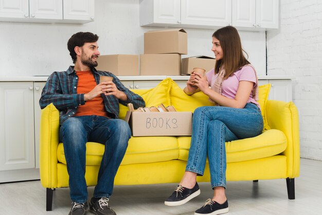 Junge Paare, die in der Hand auf dem gelben Sofa hält Tasse Kaffee Glas mit Pappschachtel im Wohnzimmer sitzen