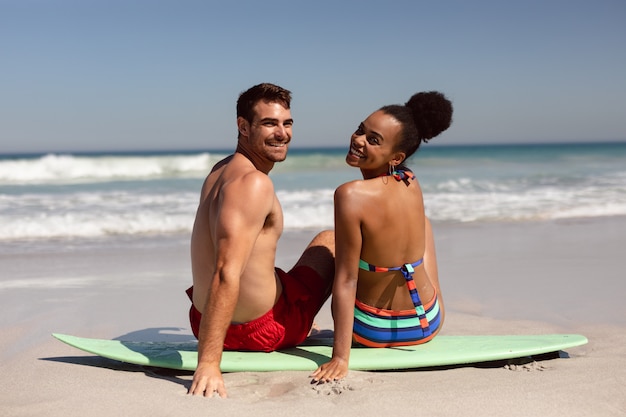 Junge Paare, die Kamera beim Sitzen auf Surfbrett am Strand im Sonnenschein betrachten