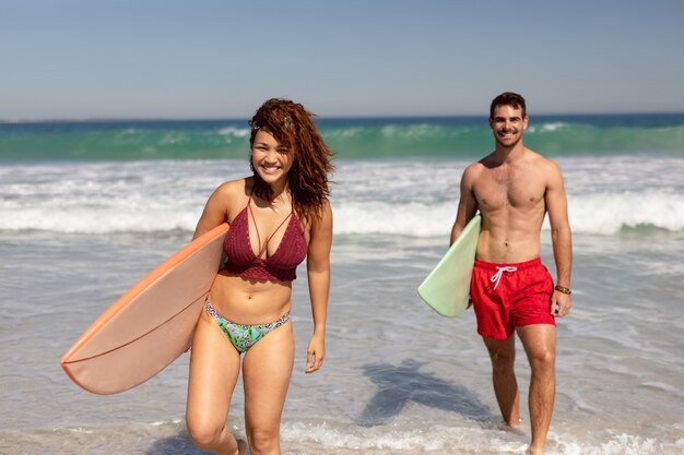 Junge Paare, die mit Surfbrett auf Strand im Sonnenschein gehen