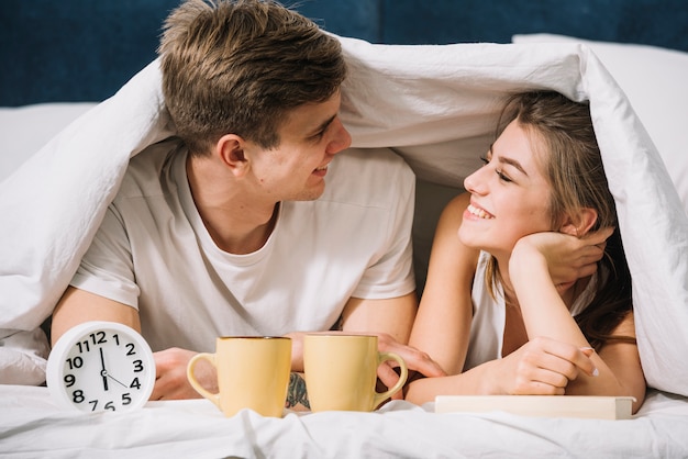 Junge Paare, die unter Decke mit Kaffee und Uhr liegen