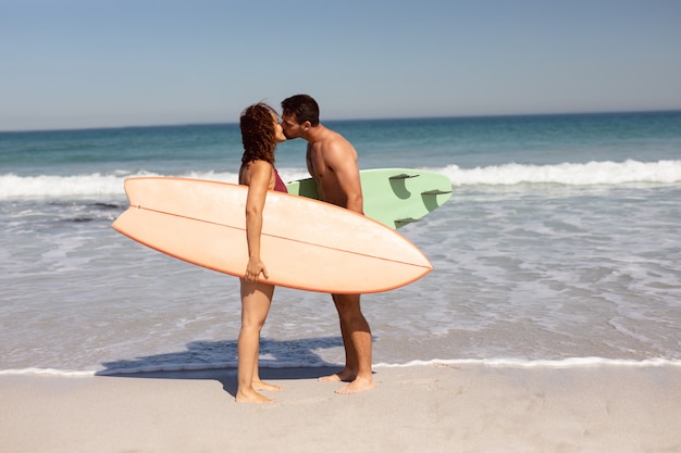Junge Paare mit dem Surfbrett, das auf Strand im Sonnenschein sich küsst