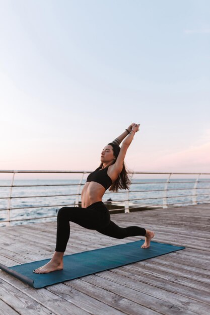Junge pansive Dame, die am Meer steht und Yoga-Posen trainiert. Schöne Frau in schwarzem, sportlichem Top und Leggings, die Yoga mit Meerblick im Hintergrund praktiziert
