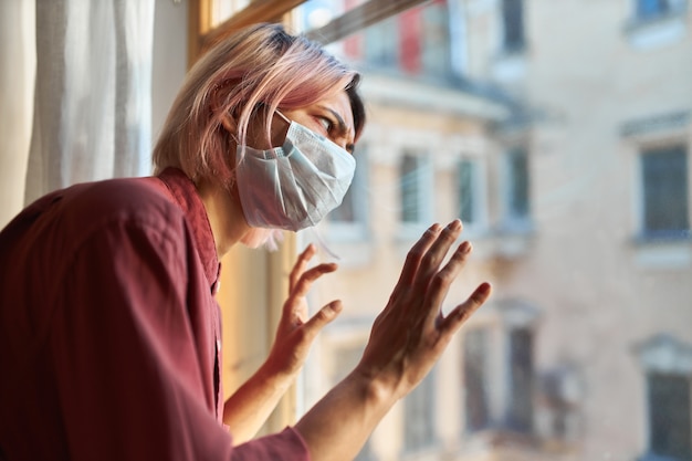 Kostenloses Foto junge patientinnen mit covid-19-symptomen müssen während der quarantäne im krankenhaus bleiben, in einer chirurgischen einwegmaske am fenster stehen, ein gestresstes paranoides aussehen haben und die hände auf glas halten