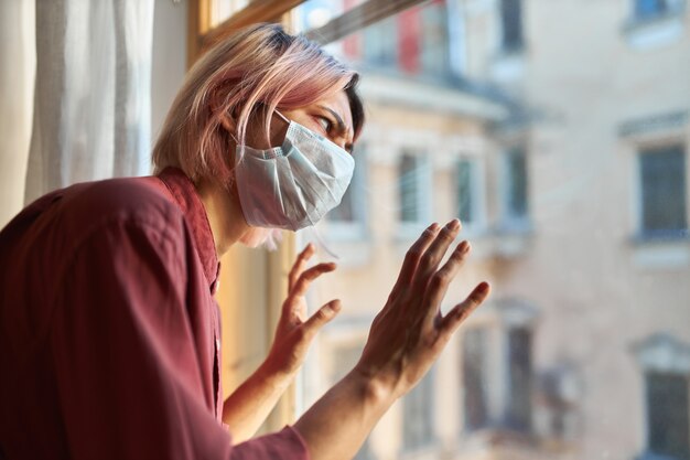 Junge Patientinnen mit Covid-19-Symptomen müssen während der Quarantäne im Krankenhaus bleiben, in einer chirurgischen Einwegmaske am Fenster stehen, ein gestresstes paranoides Aussehen haben und die Hände auf Glas halten