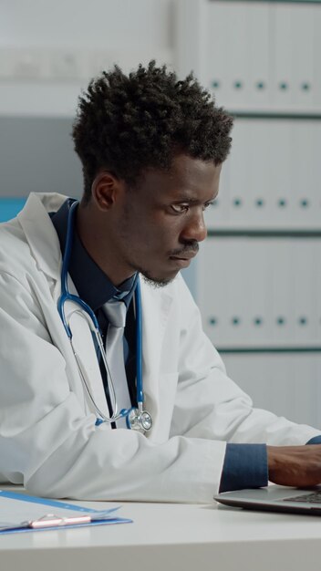 Junge Person, die als Arzt in einer Arztpraxis mit Laptop und Dokumentendateien arbeitet. Mediziner mit weißem Kittel und Stethoskop am Schreibtisch sitzend mit Technologie und Werkzeugen im Krankenzimmer