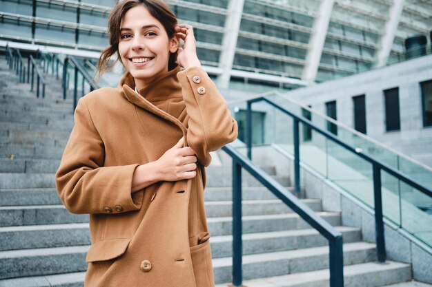 Junge positive Frau, die den Mantel vor Kälte befestigt und glücklich in die Kamera im Freien schaut